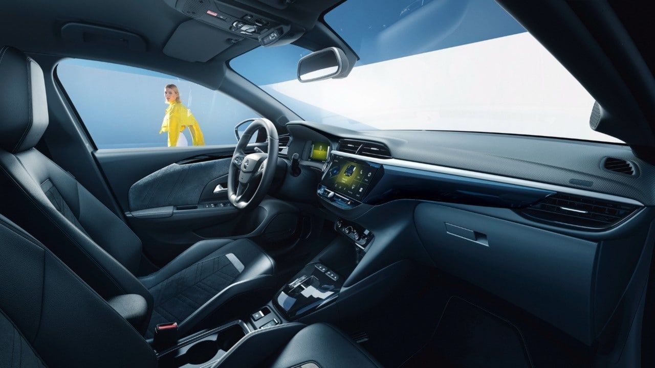 Страничен поглед на црната внатрешност кај Opel Corsa Electric од совозачкото седиште со жена во позадина во жолта кошула